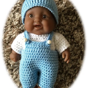 Puede incluir: Un conjunto de punto de ganchillo para una muñeca bebé, con un gorro azul, una camisa blanca y un mono azul. La muñeca tiene la piel morena y los ojos marrones.