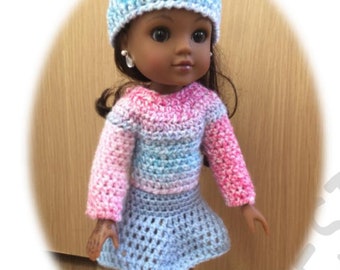 May include: A doll wearing a blue and pink striped crochet dress and a matching hat. The doll has brown skin and long brown hair.