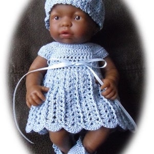 Puede incluir: Una muñeca bebé con un vestido, un gorro y botines de ganchillo de color azul claro. La muñeca tiene la piel morena y el pelo oscuro.
