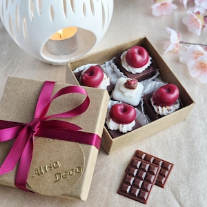 Op de afbeelding: Een bruine geschenkdoos vastgebonden met een bordeauxrode lint en een label met de tekst "Alisa Deco". De doos bevat chocoladetraktaties, waaronder vierkanten met kersen en witte crème. Twee kleine chocoladerepen zijn ook zichtbaar.