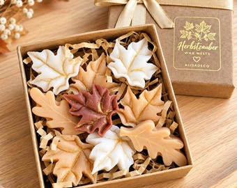 Könnte beinhalten: Eine quadratische Geschenkbox aus Karton mit blattförmigen Wachsschmelzen in Weiß, Beige und Dunkelrot, akzentuiert mit Golddetails. Der Deckel hat ein goldenes Band und ein Etikett mit der Aufschrift Herbstzauber WAX MELTS, einem Herz und ALISADECO.