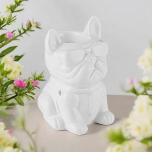 Könnte beinhalten: Eine weiße Keramik-Bulldoggen-Figur mit Sonnenbrille. Das hundeförmige Objekt hat eine ausgehöhlte Oberseite, wahrscheinlich zum Halten von Gegenständen. Die Figur ist von Blumen mit rosa und gelben Blüten umgeben.