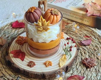 Op de afbeelding: Een gelaagde kaars in een helder glas, met pompoen- en bladvormige wasaccenten. De kaars heeft oranje, bruine en witte lagen, met een houten lont. Decoratieve herfstbladeren en een boek op de achtergrond.