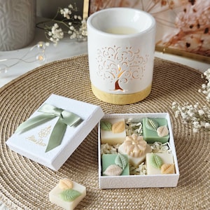 Könnte beinhalten: Ein weißer Keramik-Diffusor für ätherische Öle mit einem Baumdesign, neben einer Geschenkbox mit grünem Band und einer Schachtel mit dekorativen Seifen. Die Seifen sind quadratisch und mit Blatt- und Blumendesigns versehen.