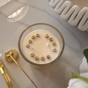 May include: A candle in a clear glass container with the words "YOUR TEXT HERE" spelled out in gold letter beads. Gold candle accessories are next to the candle. The candle has two wicks and a creamy wax color.