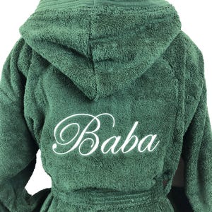 Op de afbeelding: Een donkergroene badjas met capuchon en het woord "Baba" in witte letters op de rug geborduurd. De badjas heeft een bijpassende riem en een getextureerde, zachte uitstraling, wat een comfortabel spa-gevoel suggereert.