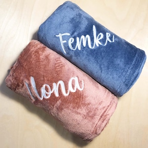 Op de afbeelding: Twee zachte, pluche dekens die naast elkaar zijn opgerold. De ene deken is stoffig blauw met de naam Femke geborduurd in wit schrift. De andere deken is gedempt roze met de naam Ilona geborduurd in wit schrift.