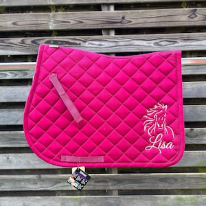 Op de afbeelding: Een felroze zadeldek voor paarden met een ruitvormig doorgestikt patroon, twee bijpassende stoffen lussen en een witte geborduurde afbeelding van een paardenhoofd boven de naam Lisa in cursief schrift, met een klein productlabel aan de onderkant.