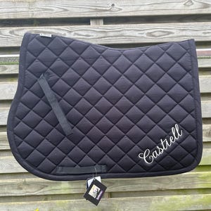 Op de afbeelding: Een zwart gewatteerd zadeldek met een ruitvormig stikselpatroon en twee zwarte nylon singelstoten. De naam Castiell is in witte cursieve letters geborduurd in de rechterbenedenhoek en er is een productlabel aan het zadeldek bevestigd.
