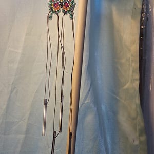 May include: A colorful butterfly wind chime with a beaded design. The butterfly has a variety of colors, including pink, yellow, green, and blue. The wind chime has several silver-colored tubes hanging from the butterfly.