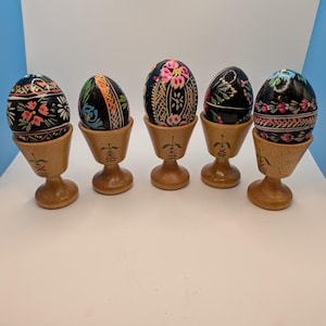 May include: Five black eggs with intricate, colorful floral and geometric hand-painted designs, each resting in a light-colored wooden egg cup. Each wooden cup features a small, delicate green and red painted floral motif on its base.