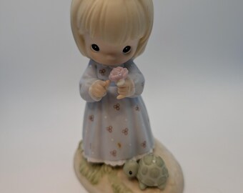 Może przedstawiać: Porcelanowa figurka blond dziecka z różową kokardą w kropki we włosach, ubranego w jasnoniebieską sukienkę w drobny kwiatowy wzór. Dziecko trzyma różowy kwiat, a u jego stóp na podstawie siedzi mały zielony żółw.