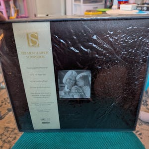 May include: A dark brown square scrapbook wrapped in clear plastic. A vertical cream-colored label on the left lists features including 12 x 12 inch page size, 20 top-loading pages, and 10 clear sheet protectors. A small square photo shows two people.