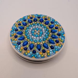 May include: A circular compact mirror featuring a decorative mandala design on the lid. The surface is embellished with a variety of blue, green, and gold rhinestones and teardrop-shaped gems arranged in a symmetrical, radial pattern.