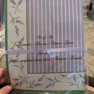 Può includere: Un set di sei rivestimenti per cassetti in una confezione di plastica trasparente. La carta presenta un bordo bianco con steli verdi e fiori di lavanda viola, e una sezione centrale con strisce verticali viola e bianche. Il testo recita Set of Six Lavender Blossom Drawer Liners.