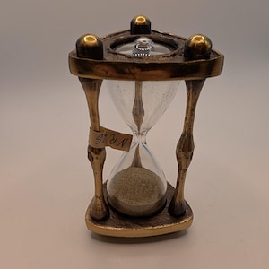 May include: An hourglass with a dark bronze-colored metal frame featuring three pillars and a triangular base. The glass bulb contains light-colored sand. A small beige adhesive label is attached to one pillar with the handwritten text NR 6.