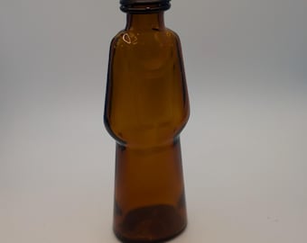 May include: A small amber-colored glass bottle with a distinct silhouette resembling a figure in a robe. The bottle features a narrow neck, rounded shoulders, and a wider base, topped with a dark brown, ribbed, screw-on metal cap.