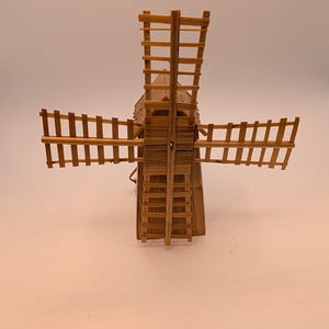 May include: A miniature windmill model constructed from light brown straw or reed material. The structure features a central tower with a pointed roof and four large, rectangular rotating blades attached to the front, all made of thin, woven slats.