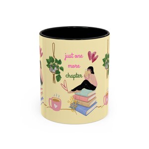 Könnte beinhalten: Ein hellgelber Keramikbecher mit schwarzem Innenrand, verziert mit einer grafischen Illustration einer lesenden Person auf einem Stapel bunter Bücher, ergänzt durch hängende Pflanzen, rosa Herzen und den Schriftzug just one more chapter.