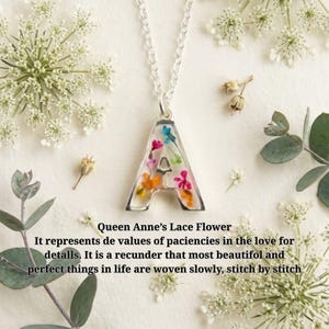 Puede incluir: Un collar de tono plateado con un colgante en forma de letra A, relleno de resina transparente e incrustado con pequeñas flores secas de colores. El colgante cuelga de una delicada cadena plateada, rodeado de flores blancas de encaje de la reina.