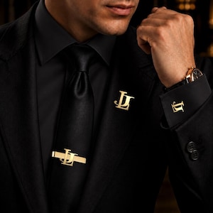 May include: A black suit jacket, black shirt, and black tie featuring a gold-toned tie clip, a gold-toned lapel pin, and gold-toned cufflinks. Each accessory displays the stylized gold letters JL in a matching font and metallic finish.