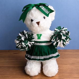 Könnte beinhalten: Ein weißer Plüsch-Teddybär als Cheerleader verkleidet, mit einer grün-weißen Uniform mit einem Buchstaben V auf der Brust, einem grünen Rock mit silbernem Saum und einer grünen Haarschleife mit dem Namen Jamie in silbernem Glitzer, hält zwei Pompons.