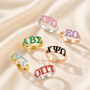 May include: A collection of six metal rings featuring Greek letters in various enamel colors including pink, green, purple, black, light blue, and red. Each ring displays a unique combination of three Greek characters set on a thin metallic band.