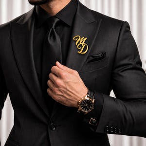 May include: A black suit jacket with a matching black shirt and tie. A gold-toned metal brooch featuring the stylized letters MD is pinned to the left lapel. The jacket features a pocket square, and the sleeve cuffs have black square buttons.