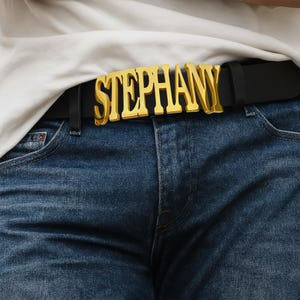 Pode incluir: Um cinto de couro preto com uma fivela de metal grande na cor dourada que forma o nome STEPHANY em uma fonte em negrito. O cinto é usado através dos passantes de uma calça jeans azul, posicionado abaixo de uma camiseta branca lisa.