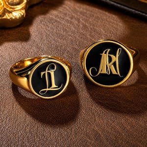 May include: Two gold-toned rings with circular faces featuring black enamel inlays. The smaller ring displays a gold letter L in a cursive script, while the larger ring features a gold monogram of the letters AR in a matching cursive script.
