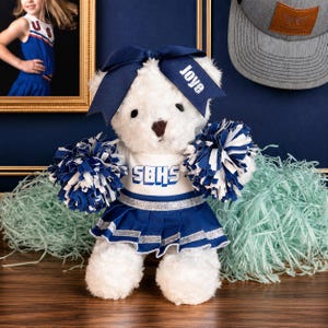 May include: A white plush teddy bear dressed as a cheerleader, wearing a navy blue and white pleated skirt and top with the text SBHS. The bear has a navy blue hair bow with the name Joye and holds two blue and white pom-poms.