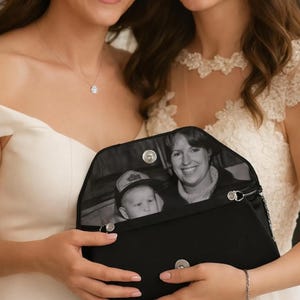 Peut inclure: Pochette noire avec une chaîne argentée. La pochette est ouverte, révélant une photographie en noir et blanc d'une femme et d'un enfant. La pochette est tenue par deux personnes, dont une portant une robe blanche.