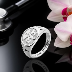 May include: A silver ring with a medical symbol and the text "199" and "Love and care." The ring has a polished finish and is set against a black background with a stethoscope and flowers.