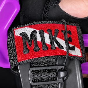 May include: A black fabric strap featuring a rectangular patch with a red background, a white diagonal stripe, and the name MIKE embroidered in black thread. The strap includes a black plastic buckle and a thin black cord with a sliding toggle.