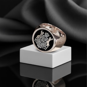 May include: A rose gold signet ring with a black face. The face features a detailed crest with deer, a crown, and the name "O'Connell". The ring is displayed on a white cube against a dark background.