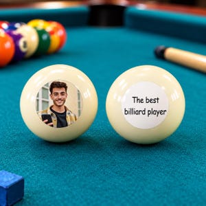 May include: Two off-white billiard balls resting on a green felt table. One ball features a circular colour photograph of a smiling person holding a smartphone. The other ball displays the black printed text, The best billiard player.