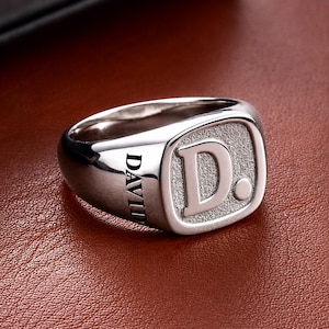 May include: A polished silver-tone signet ring featuring a square face with a textured background and the letter D followed by a full stop. The side of the ring band is engraved with the name DAVID in a clean, sans-serif capital letter font.