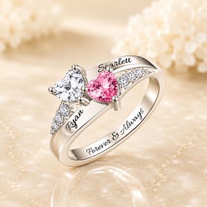 Può includere: Un anello color argento con due pietre a forma di cuore, una trasparente e una rosa, affiancate da piccole pietre trasparenti. La fascia è incisa con i nomi Ryan e Scarlett e la frase Forever & Always, con una finitura metallica lucida.
