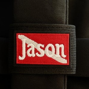Puede incluir: Un parche de tela negra con el nombre "Jason" bordado en blanco sobre un fondo rojo. El parche rectangular está adherido a una correa negra, probablemente parte de una bolsa o accesorio.