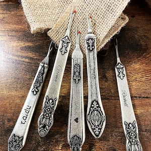 May include: Five silver seam rippers with decorative patterns and red tips are shown on a wooden surface. Each ripper has a different name engraved on it. A burlap cloth is in the background.