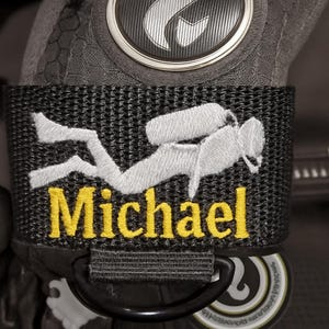 Op de afbeelding: Close-up van een zwart en grijs duikaccessoire. Een zwarte band heeft een wit geborduurd duikersilhouet en de naam "Michael" in geel. Het accessoire heeft een zilveren embleem en een gestructureerd oppervlak.