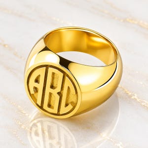 May include: A polished gold-tone signet ring featuring a circular face with the letters A, B, and C engraved in a bold, block-style monogram. The ring has a smooth, rounded band and a high-shine metallic finish that reflects light across its surface.