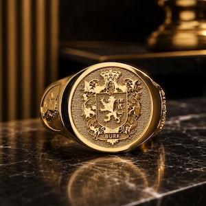 May include: A gold signet ring featuring an oval face with an embossed heraldic crest. The design includes a central lion figure, ornate scrollwork, a crown at the top, and the name BURK inscribed in capital letters at the base of the shield.