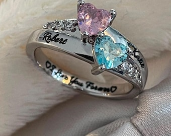 May include: A silver-toned ring featuring two heart-shaped stones, one pink and one light blue, set side-by-side. The band is engraved with the names Robert and Katie, and the inner surface displays the engraved text I Love You Forever.