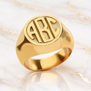May include: A polished gold signet ring featuring a circular face with the engraved letters ABC in a block font. The ring has a smooth, reflective surface and a thick band, showcasing a classic design with a textured background behind the letters.