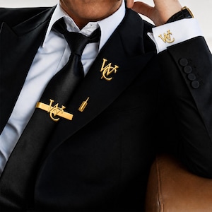 May include: A black suit jacket with a white dress shirt and a black satin tie. The set includes a gold tie bar, a gold lapel pin, and gold cufflinks, all featuring the gold monogram letters WC in a stylized, interconnected script font design.