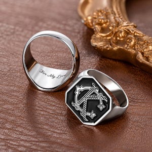 May include: Two silver-toned rings. One ring features a flat, octagonal face with a black inlay and a prominent letter K design. The second ring is positioned to show the interior band, which is engraved with the cursive text For My Love.