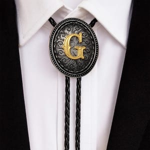 Può includere: Cravatta a bolo ovale con cornice argentata e lettera "G" dorata. La cravatta ha un cordino nero intrecciato e viene indossata su una camicia bianca e un abito nero. Accessorio in stile western.
