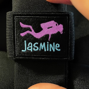 Può includere: Toppa in tessuto nero con un disegno di subacqueo viola e il nome "Jasmine" ricamato in turchese. La toppa è rettangolare con un bordo nero, fissata a un materiale nero strutturato.