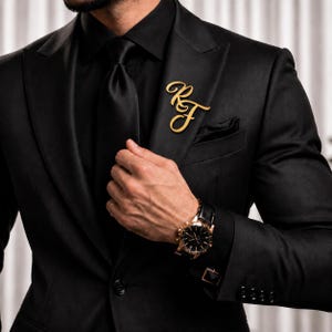 Può includere: Una giacca da abito nera con camicia e cravatta nere abbinate, caratterizzata da una spilla da bavero in metallo dorato a forma di lettere corsive RF. La giacca presenta un bavero dentellato, un fazzoletto da taschino e un orologio nero.
